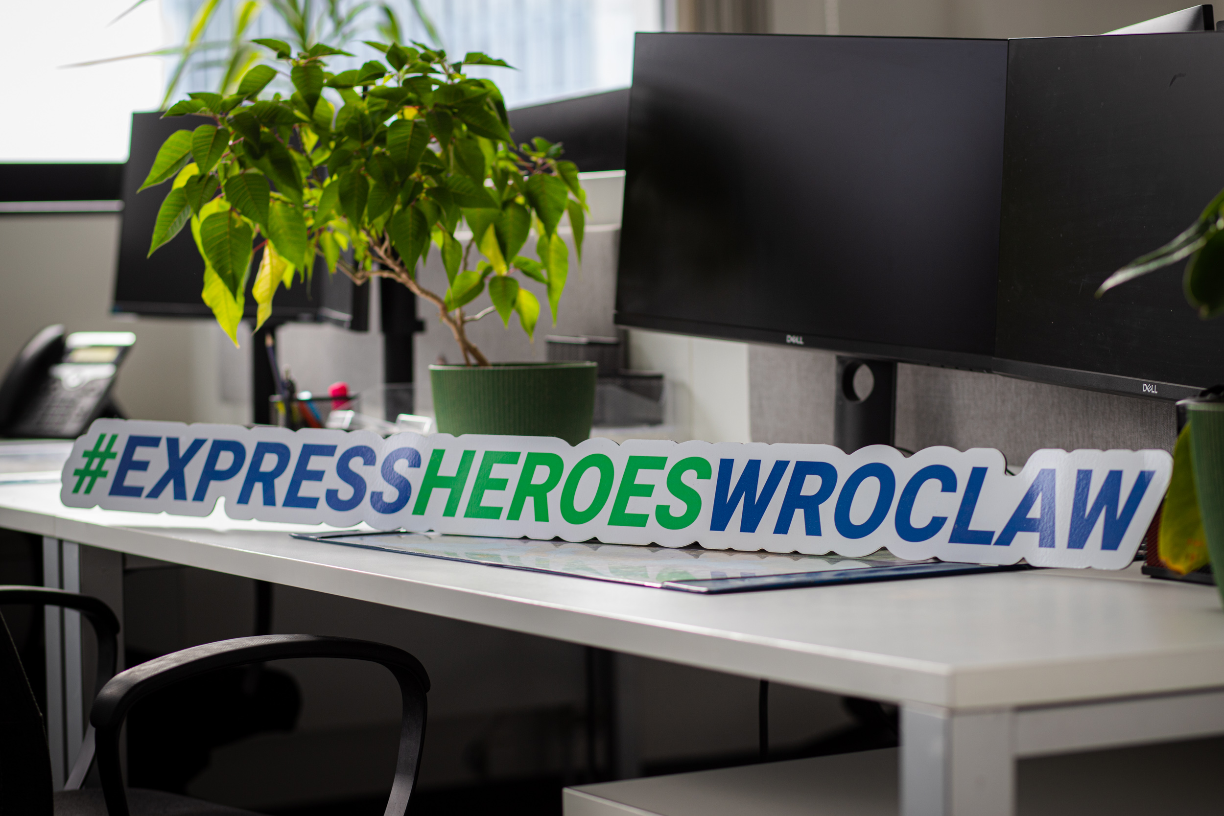 Desk with monitors, a potted plant and a #ExpressHeroesWroclaw sign at the Express Heroes office.
