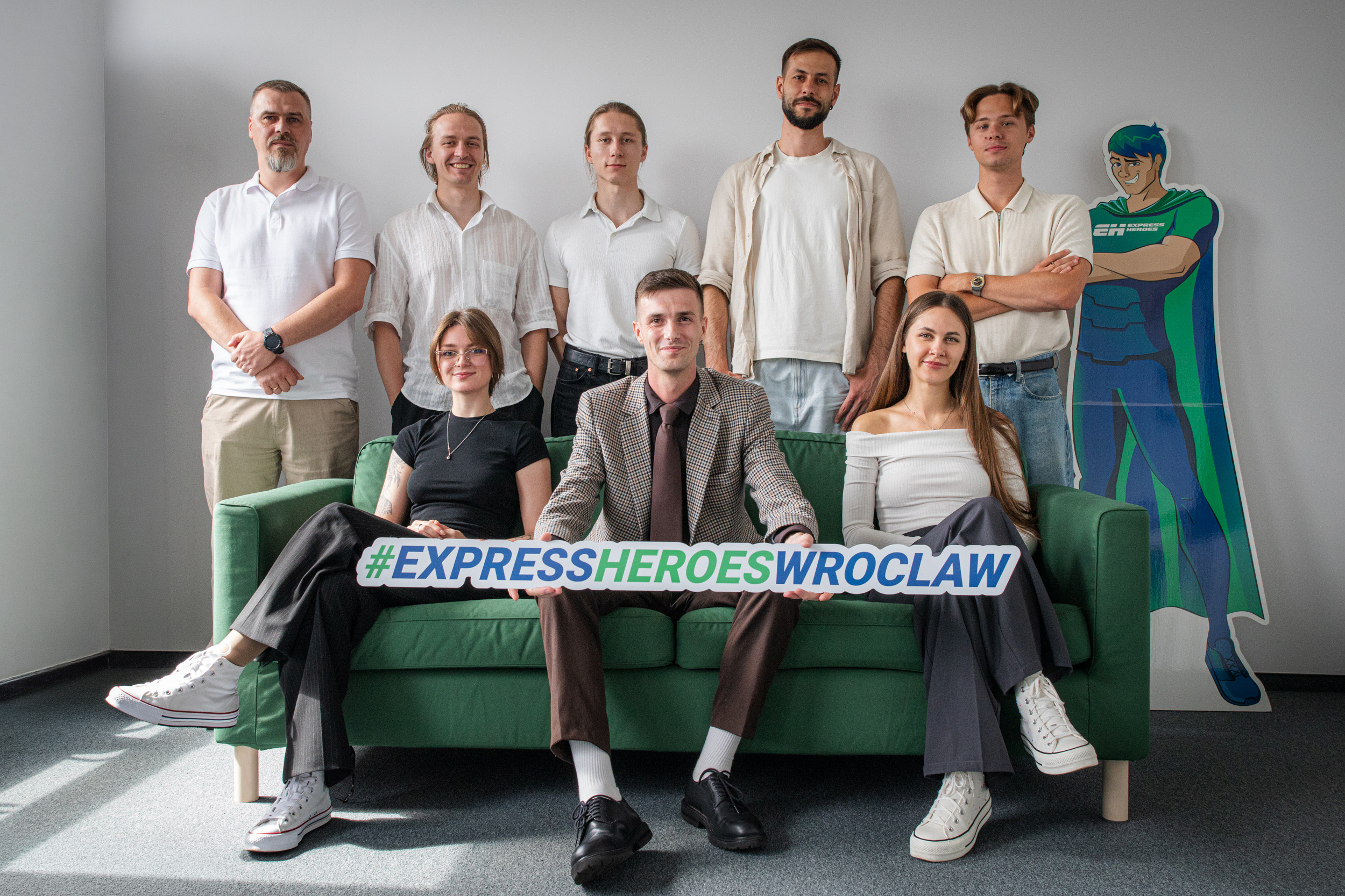 Express Heroes Wrocław team gathered around a green sofa, holding a sign with the office hashtag.
